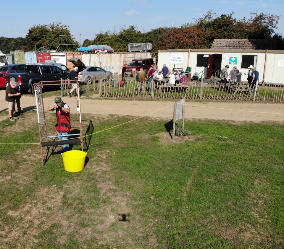 A clay shooting event at Fenland Gun Club in the UK. A shooter wearing safety gear and ear protection is aiming a shotgun from a marked shooting stand, with a yellow bucket in front. Several people are gathered near the clubhouse in the background, while others stand by parked cars. The scene takes place on a sunny day with clear skies.