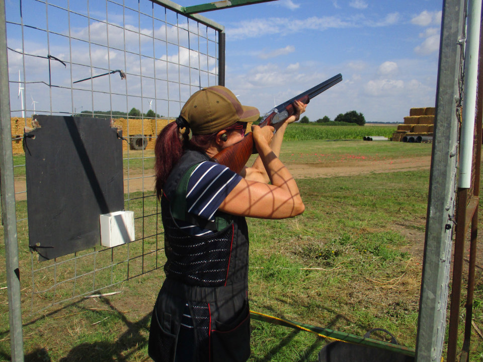 A woman participating in clay shooting at Fenland Gun Club in the UK. She is wearing a cap, ear protection, and a shooting vest, aiming a shotgun from within a designated shooting stand. The background shows fields, hay bales, and wind turbines under a partly cloudy sky.